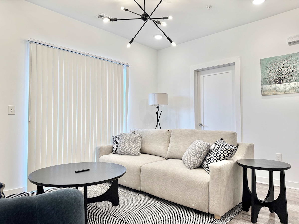 A light-filled living area features a comfortable, neutral-toned sofa adorned with decorative pillows. Two round black tables are positioned nearby, complemented by a modern light fixture overhead. Vertical blinds allow natural light to filter in, enhancing the open atmosphere of the space.