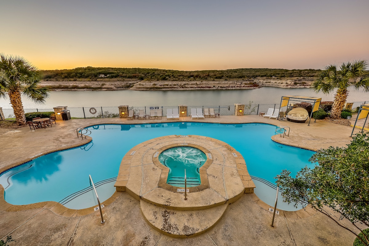 A calm pool area is visible, featuring a large circular hot tub and a spacious swimming pool. Surrounding palm trees offer a tropical feel, while lounge chairs line the pool deck. The tranquil waters of the lake can be seen in the background, reflecting the colors of dusk.