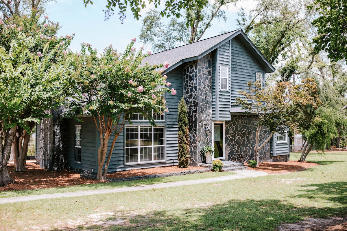 A spacious two-story home features a stone facade and blue siding, surrounded by lush trees and manicured lawns. Large windows allow for ample natural light, while a path leads to the entrance. Soft landscaping elements add a touch of greenery to the outdoor space.