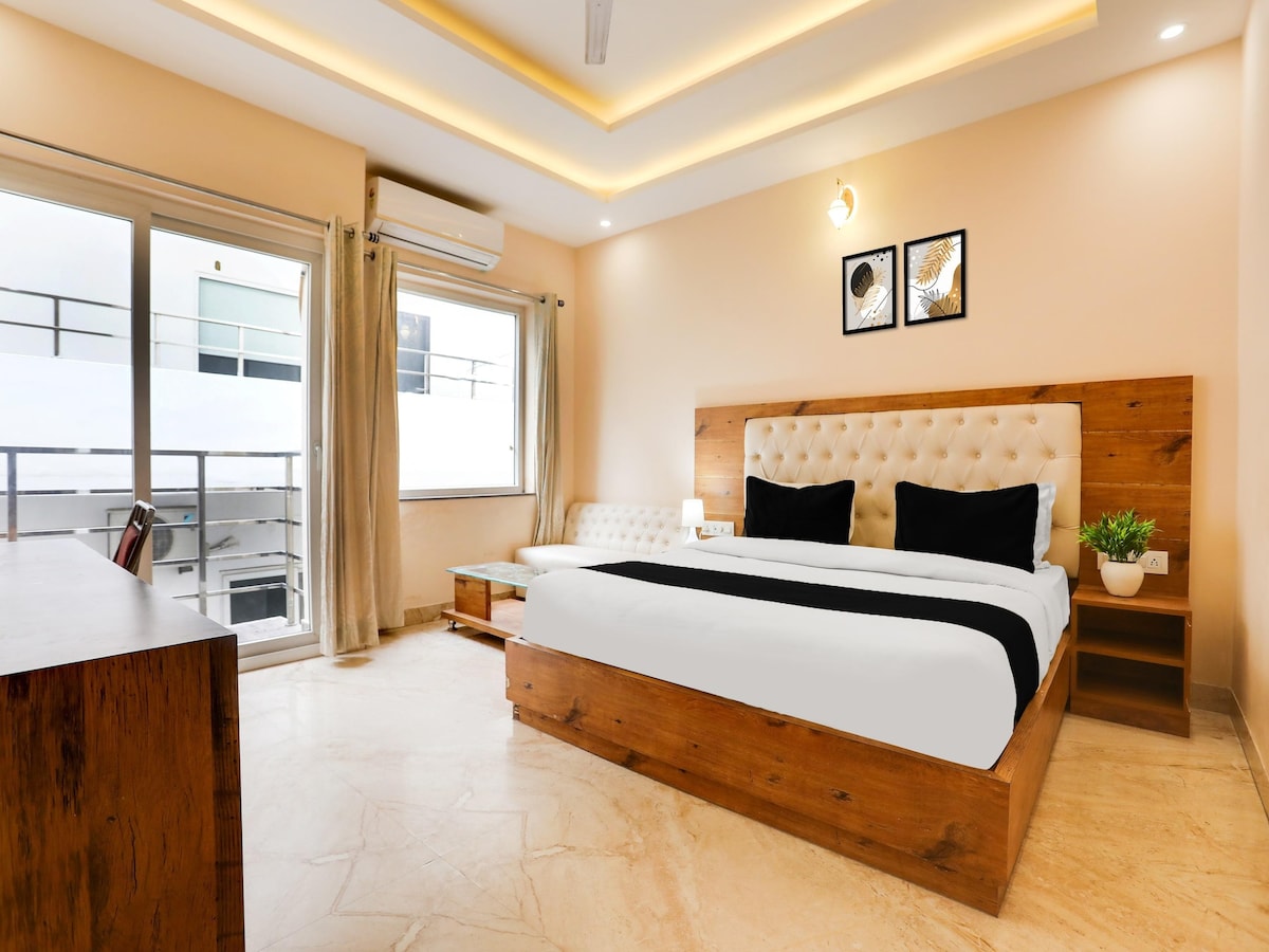 A bright guest room is furnished with a spacious wooden bed featuring an upholstered headboard. A comfortable seating area is visible beside a large window, providing natural light. A desk and a small plant adorn the room, with framed art displayed on the wall.