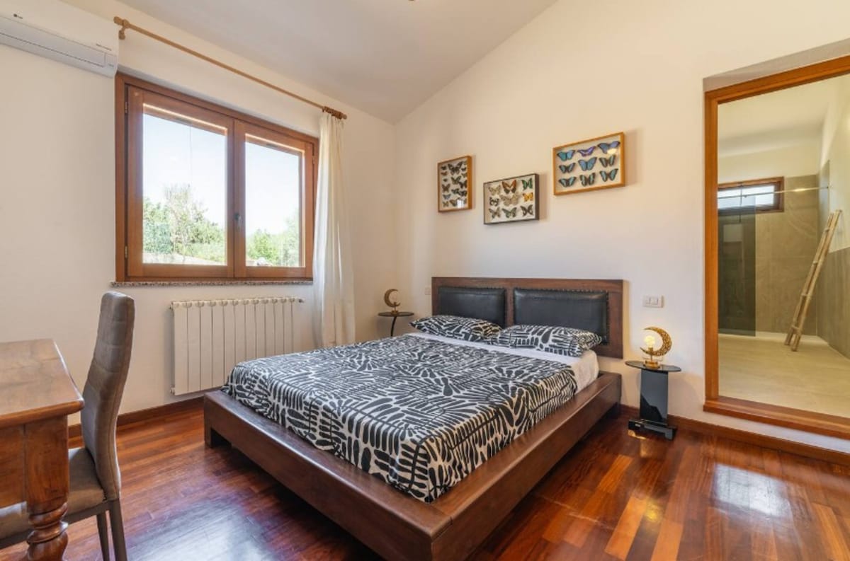 A bright bedroom is presented, featuring a spacious bed with a patterned duvet cover. Natural light filters through two large windows, illuminating the space. Wall art depicting butterflies adorns the walls, alongside a wooden desk. The adjoining bathroom area is visible in the background.