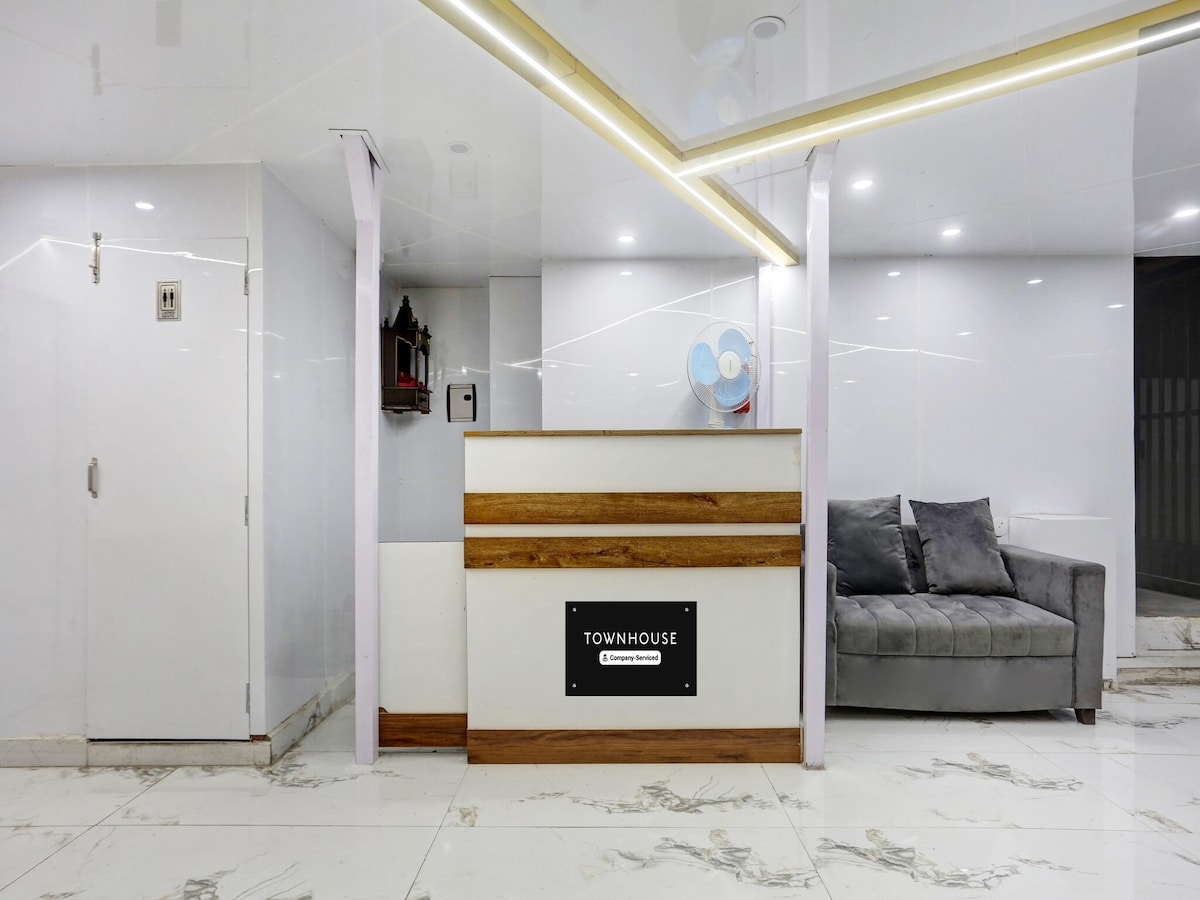 A reception area featuring a sleek, modern desk with wooden accents. A comfortable gray seating area is positioned nearby, and bright white walls enhance the clean aesthetic. Decorative elements and a fan are subtly included, contributing to the minimalist design.