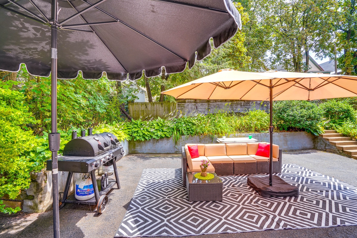 A shared backyard area is presented, featuring a charcoal grill beside a sectional seating arrangement covered by an umbrella. A patterned rug anchors the space, surrounded by lush greenery and a stone wall. A second umbrella provides additional shade over the seating.