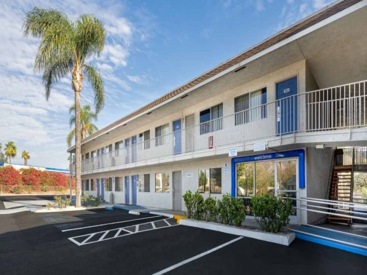 The exterior of the two-story building is shown, featuring a welcoming entrance with blue doors and railings. Palm trees are positioned nearby, and a well-maintained parking area is prominently visible in the foreground, accompanied by landscaped greenery.
