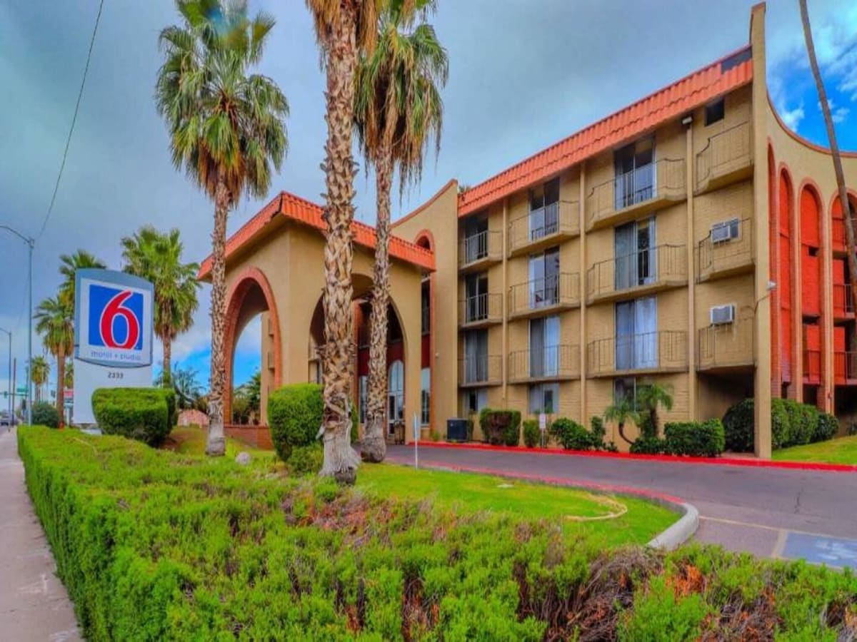 The exterior of the hotel is depicted, showcasing a multi-story building with a distinctive architectural style. Palm trees are featured prominently in the foreground, alongside neatly trimmed shrubs. A clear sky is visible above, and the motel's signage is positioned nearby.