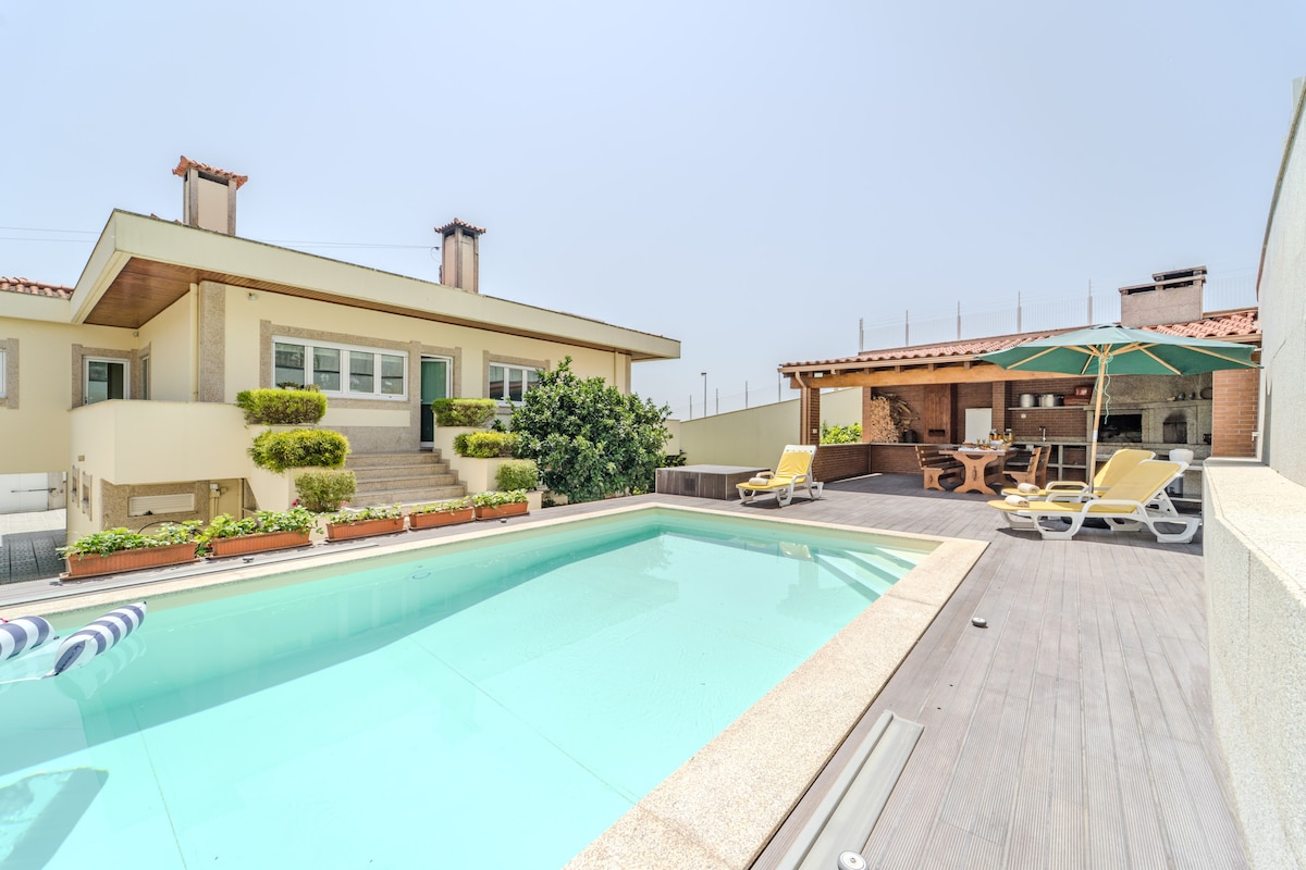 A well-maintained outdoor area features a private swimming pool with clear water. Surrounding the pool are sun loungers arranged for relaxation. A cozy barbecue area with outdoor dining furniture and an umbrella is visible, along with lush green plants bordering the space.