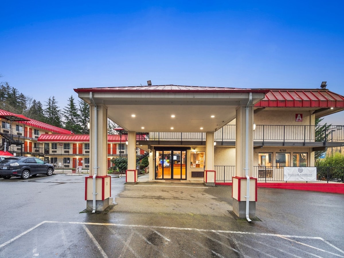 The entrance to the hotel is highlighted by a covered portico, featuring overhead lighting. The surrounding area includes parking spaces and a glimpse of the hotel’s rooms, which have vibrant red roofs. Lush trees are visible in the background.