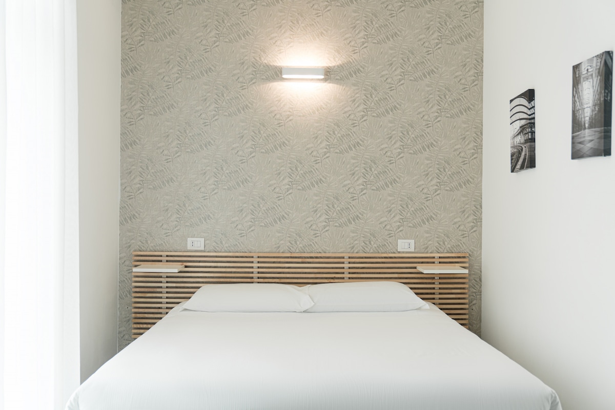 A modern studio sleeping area features a spacious bed with a simple white duvet against a patterned wallpaper backdrop. Two black-and-white photographs adorn the wall, while soft lighting highlights the bed's wooden headboard. Natural light filters through sheer curtains near the window.
