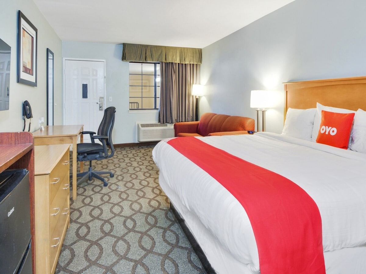 A spacious room features a king bed with crisp white bedding and a bright OYO pillow. A desk and office chair are positioned near the entrance, while a seating area with a red sofa is visible. Large windows allow natural light to fill the space.