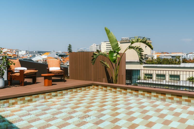 A rooftop pool area features patterned tiles in a light blue and beige design. Two lounge chairs are positioned nearby, each with a towel neatly placed on the seat. A banana plant adds greenery to the space, with a clear sky and city views in the background.