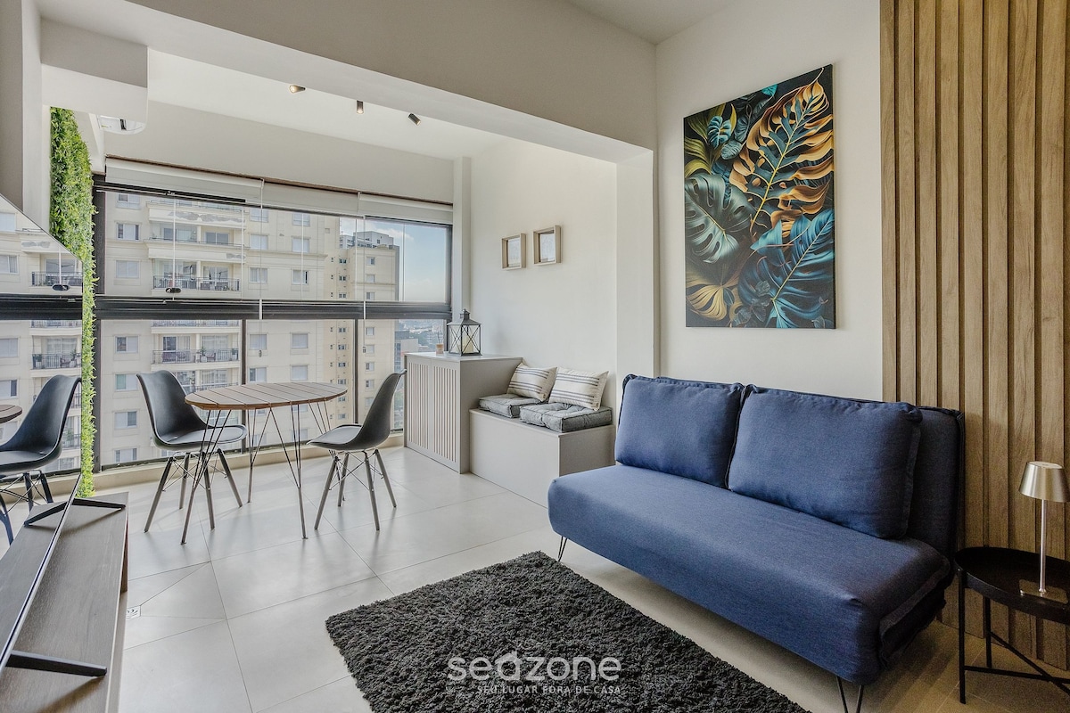 The living area features a comfortable sofa bed and a small dining table with chairs. A plush dark rug is underfoot, and natural light fills the space from large windows, offering a view of the city. A vibrant wall art piece adds a touch of color.