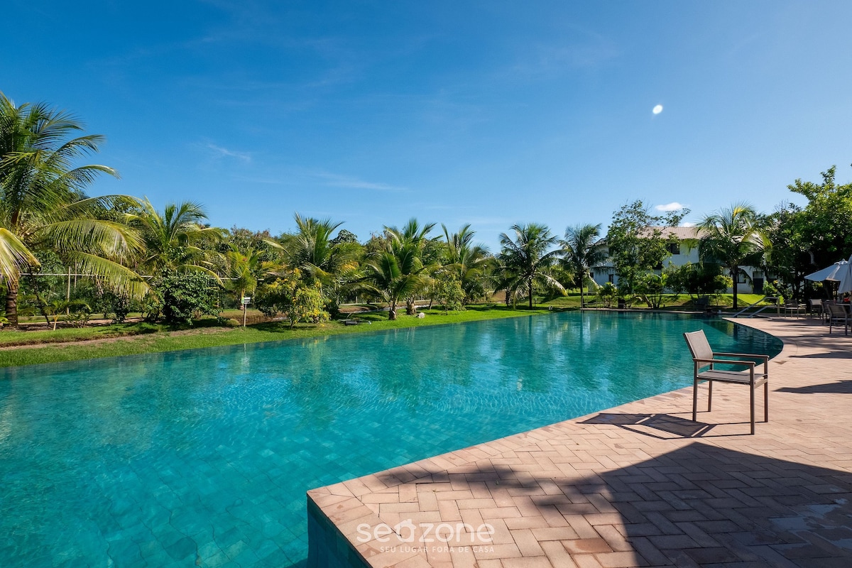 A serene swimming pool is situated amidst lush greenery, featuring clear turquoise water that reflects the sky. Palm trees and landscaped gardens surround the area, creating a tranquil environment. A single chair is positioned nearby on the deck, inviting relaxation.