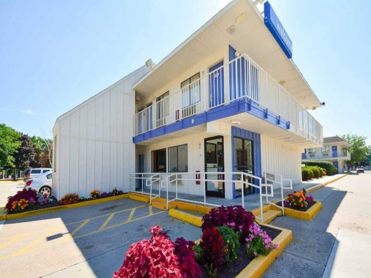 The exterior of a motel features a white facade with blue accents and a wide staircase leading to the upper level. Colorful flowerbeds with vibrant blooms add a decorative touch along the entrance pathway, enhancing the inviting appearance of the property.