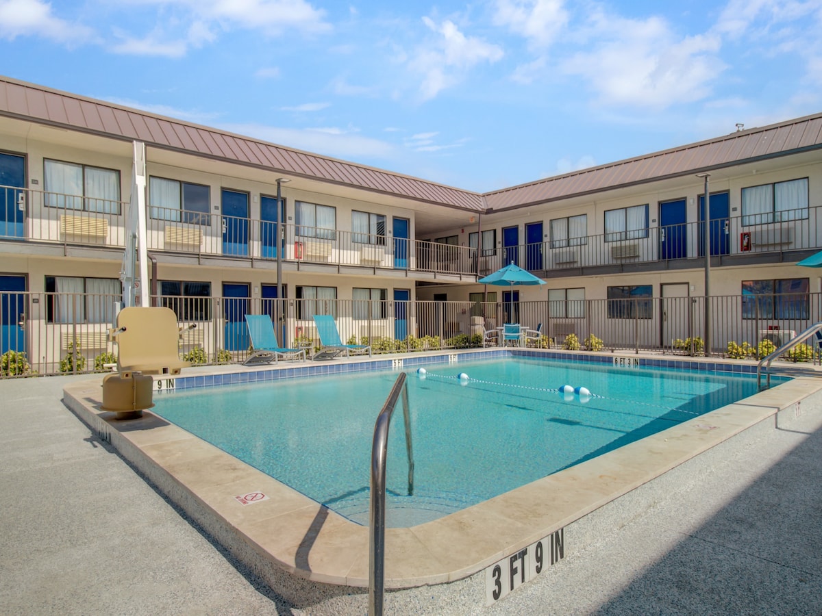 An outdoor swimming pool is centrally located within a courtyard setting, surrounded by sun loungers and umbrellas. The pool area features clear water and a depth marker at one end. Guestroom balconies are visible behind the pool, providing a view of the surrounding space.