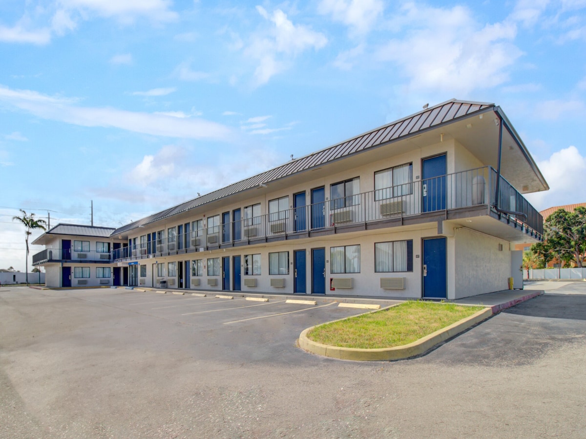 The exterior of the motel features a two-story building with a beige façade and blue doors. Balconies can be seen on the upper level, while the parking area is spacious and well-maintained, allowing for easy access. A clear blue sky enhances the overall appearance.