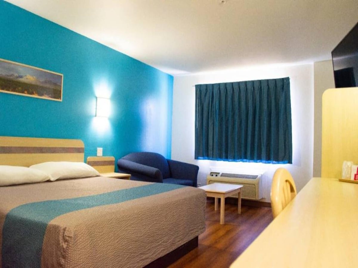A comfortable room is showcased, featuring a spacious king bed with a light grey coverlet. A cozy sofa and small table are positioned beside a window dressed in dark blue curtains. A flat-screen TV is mounted on the wall, with a wooden desk and chair located nearby.