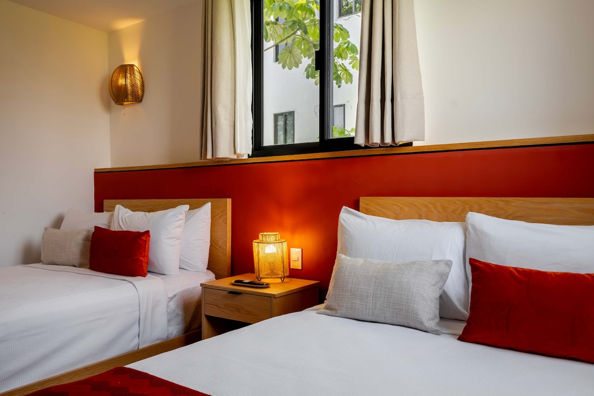 A cozy sleeping area features two spacious beds with soft white bedding and decorative pillows in warm tones. A bedside lamp softly illuminates a wooden nightstand. Natural light filters through a large window, complemented by light drapes, creating a calm environment.