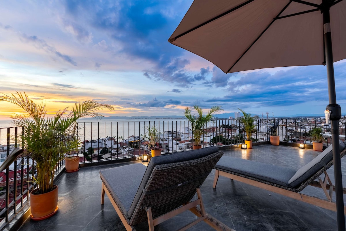 A spacious rooftop terrace is presented, featuring lounge chairs and decorative potted plants. The area is framed by a stylish railing, offering expansive views of the ocean and surrounding city. Warm hues from the sunset create a calming backdrop, enhancing the inviting outdoor space.