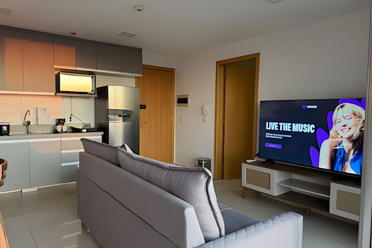 A modern kitchen is visible, featuring sleek cabinetry and stainless steel appliances. A comfortable gray sofa is positioned in front of a large Smart TV. Natural light fills the space, highlighting the open layout and contemporary design elements of the apartment.