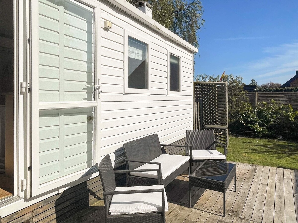 A wooden deck is shown, featuring outdoor furniture including two chairs and a small table. The bright exterior of the cottage is partially visible, with large glass doors providing access to the interior. Surrounding greenery adds a natural touch to the setting.