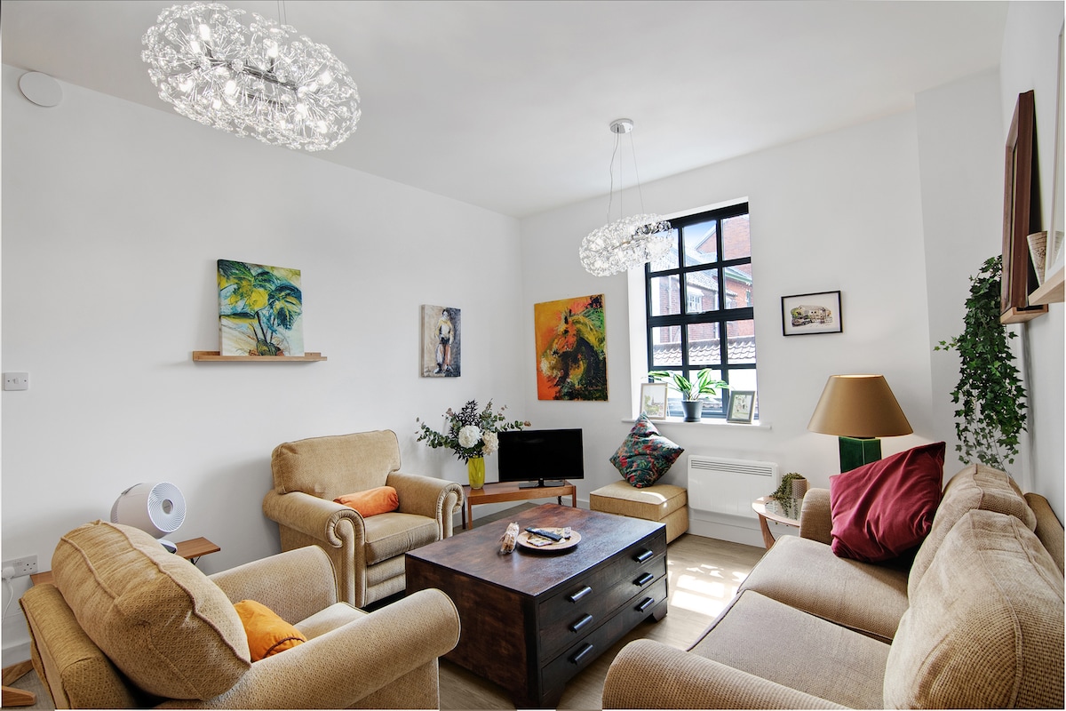 The living area is furnished with comfortable seating, including two armchairs and a sofa, arranged around a wooden coffee table. Original artwork adorns the walls, and large windows allow natural light to fill the space. Contemporary light fixtures hang from the ceiling, enhancing the room's ambiance.
