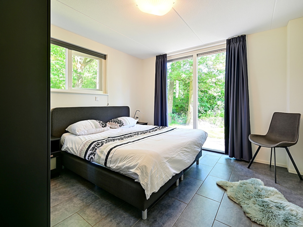 A spacious bedroom features a comfortable bed dressed in white linens with dark accents. Large windows allow natural light to fill the room, providing views of the greenery outside. A chair sits near the window, and soft textures are present on the floor.