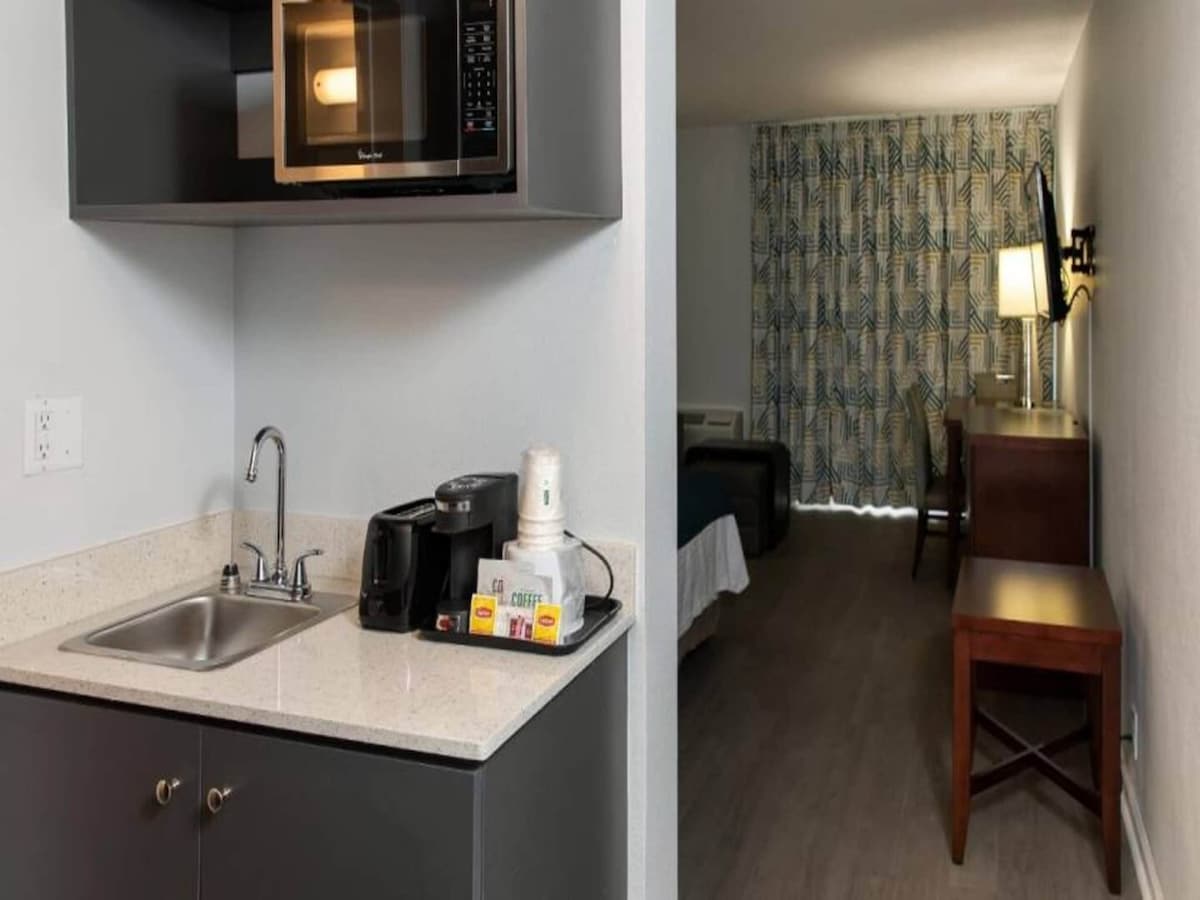 A kitchenette is featured with a sink, microwave, and coffee maker. Packaged coffee supplies and a small selection of tea are visible. The area is contrasted by warm wood tones from the cabinetry and a desk situated in the background, with a patterned curtain visible.