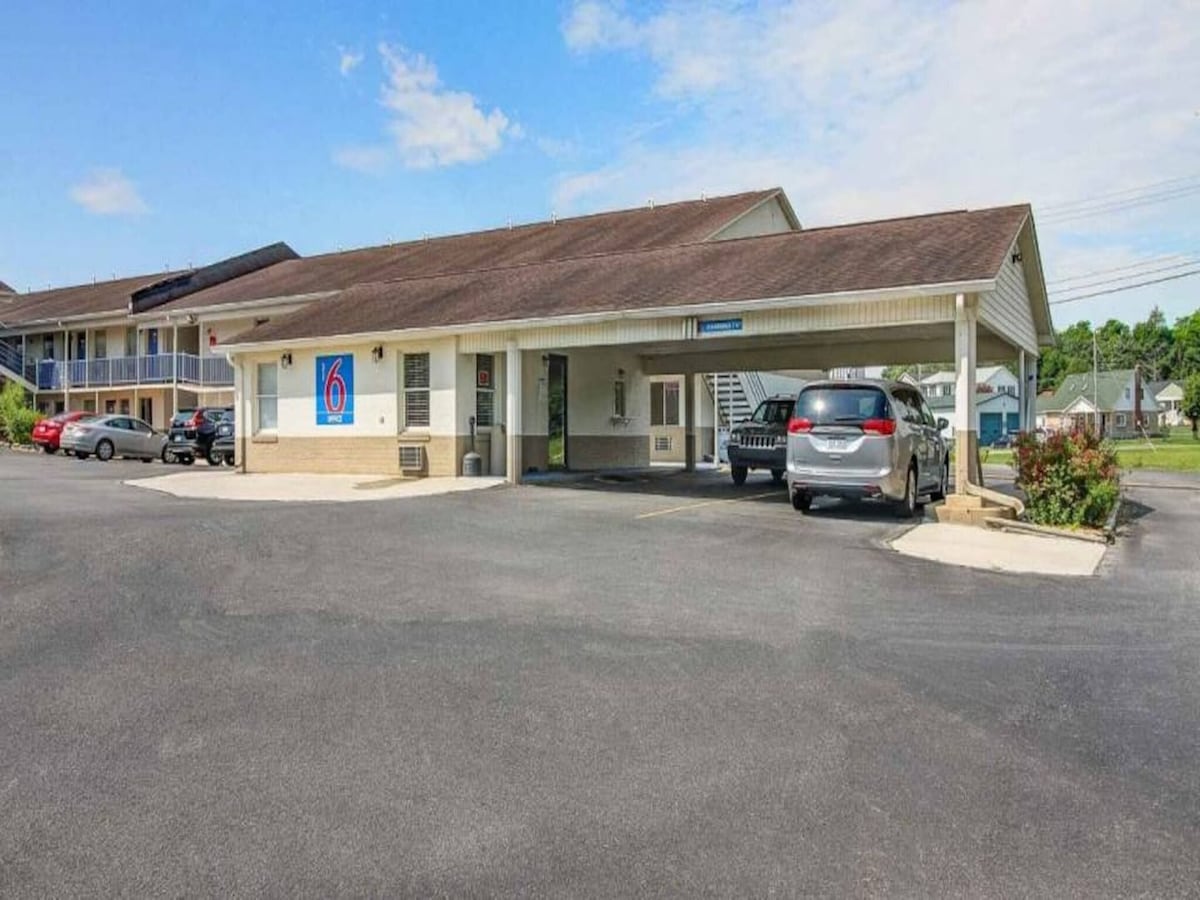 The exterior of the Motel 6 features a single-story structure with a light-colored facade and a prominent sign. A paved parking area can be seen with several vehicles parked. Green landscaping and clear blue skies provide a welcoming environment.