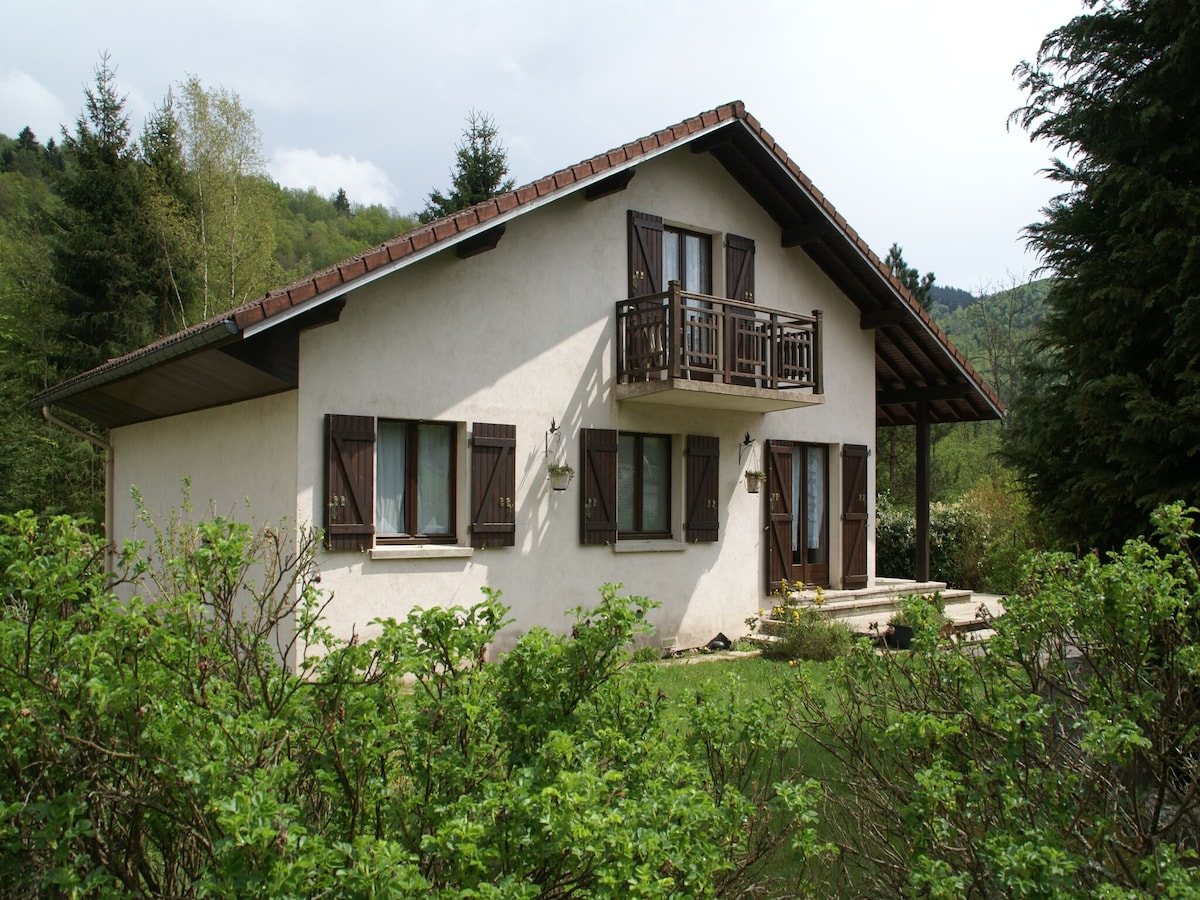A two-story chalet is nestled among greenery, showcasing a light-colored facade. Multiple windows and wooden shutters are present, with a balcony above the entrance. The sloped roof and surrounding trees enhance the natural setting of the property.
