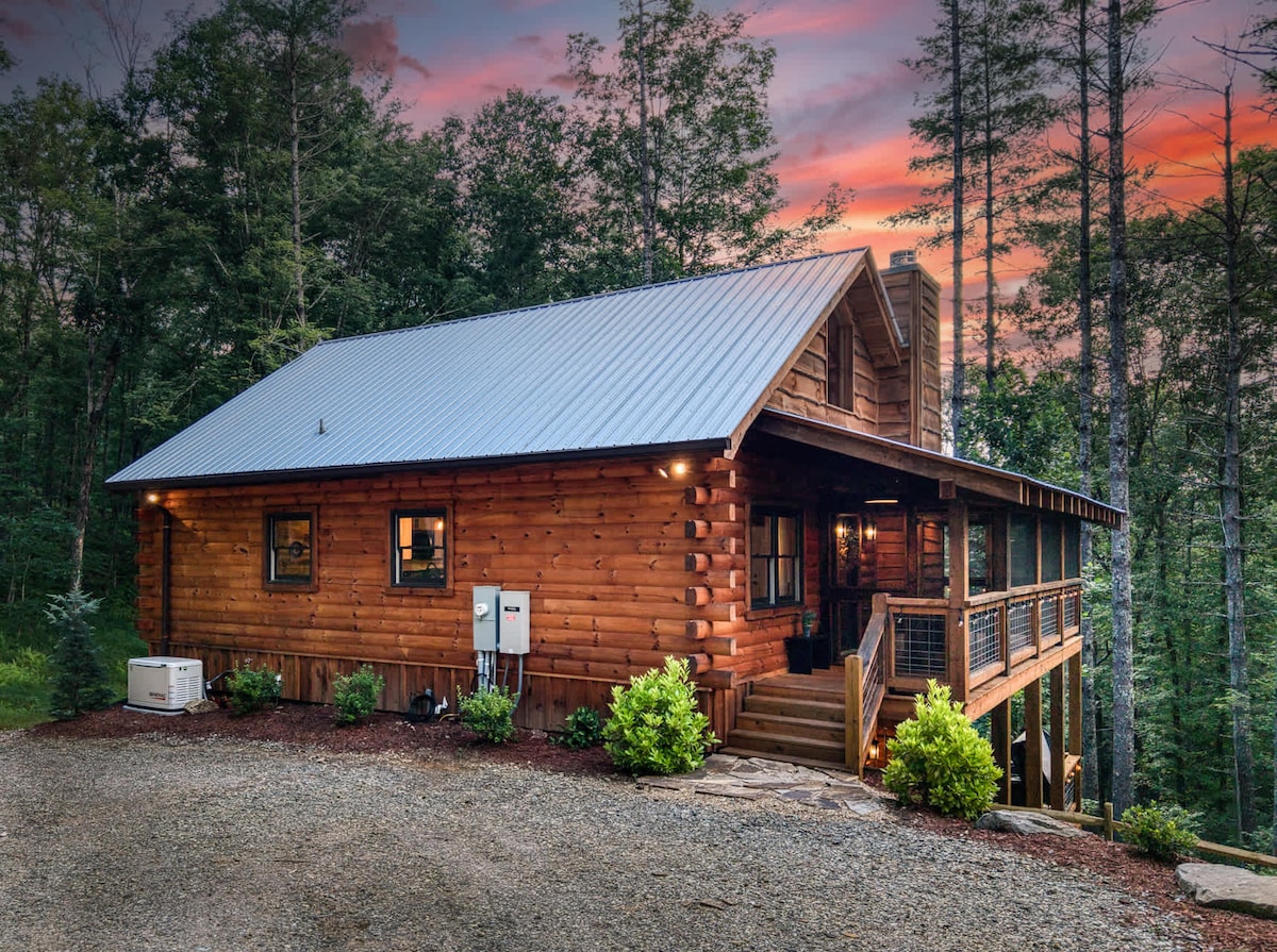 A charming log cabin is depicted, featuring a metal roof and large front porch. Surrounded by trees, the home exhibits a warm glow from exterior lights. A gravel driveway leads up to the property, and vibrant green bushes are positioned near the entrance.