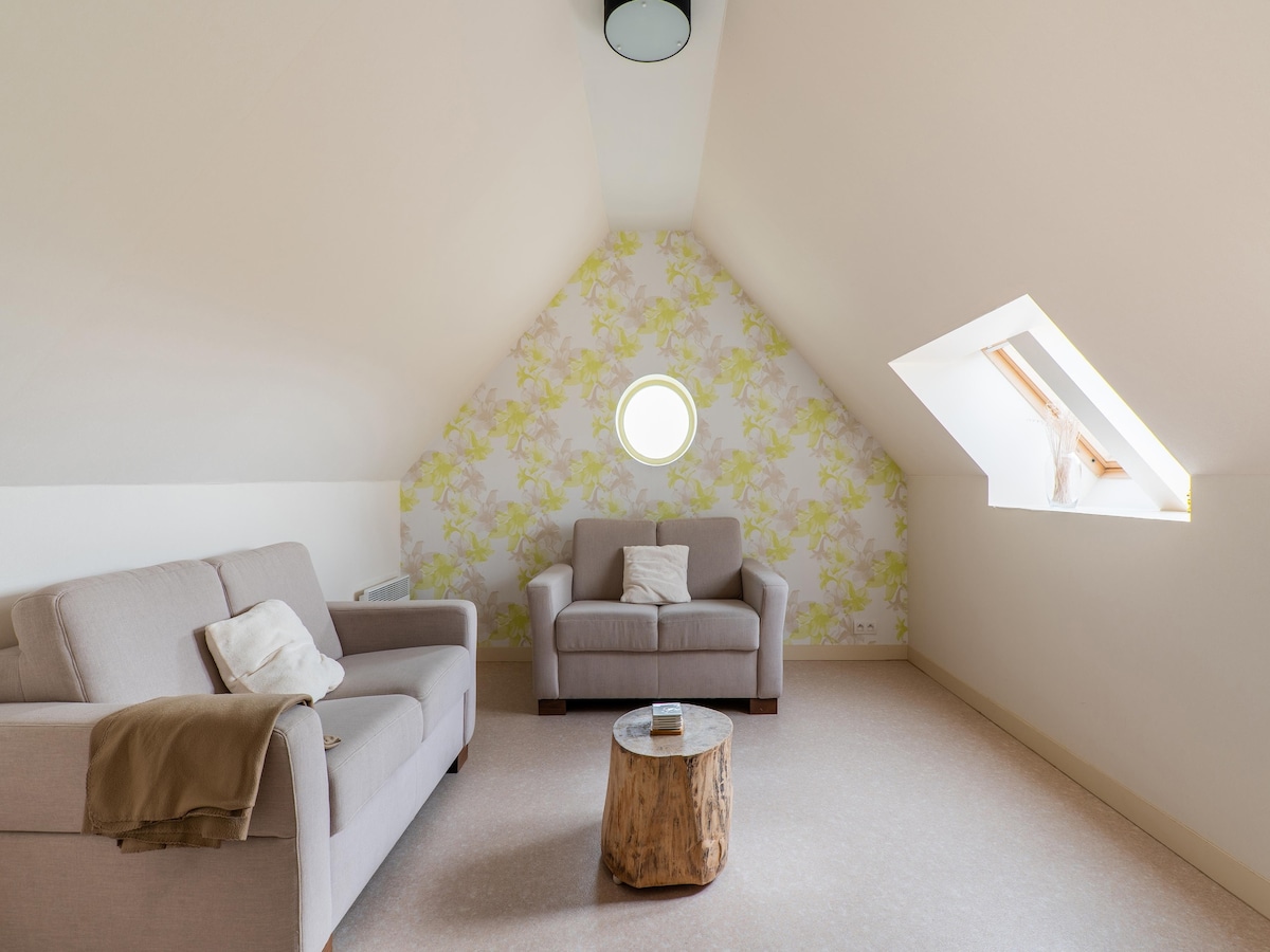 An inviting living space features two light-colored sofas arranged around a natural wood coffee table. A circular window allows natural light to flood the room, while a floral accent wall adds a subtle touch of color. The sloped ceiling contributes to a cozy atmosphere.