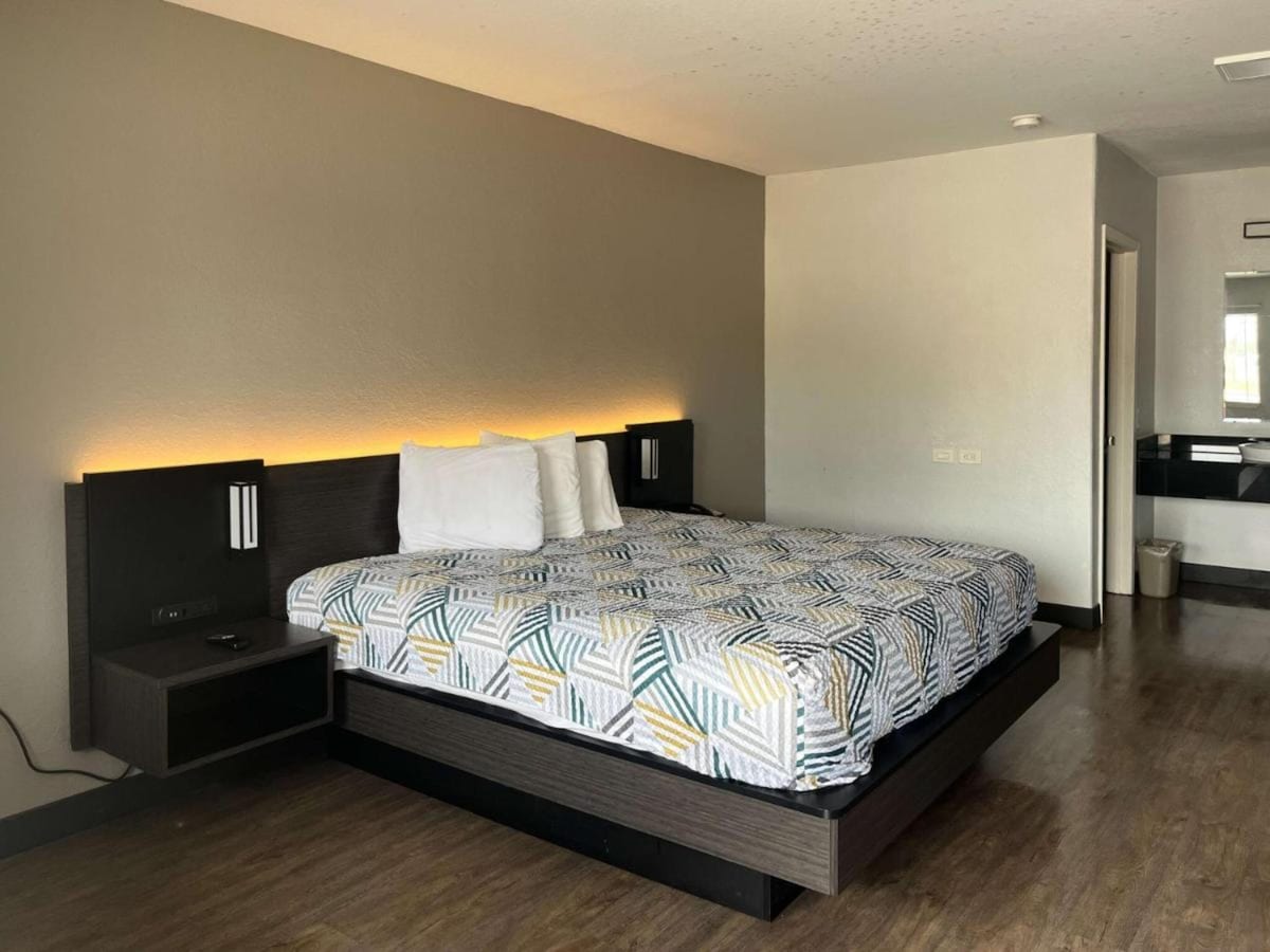 The room features a spacious king bed adorned with a patterned duvet and white accent pillows. Soft lighting is positioned on the headboard. A minimalist nightstand is placed beside the bed, and an open area leads to a bathroom visible in the background.