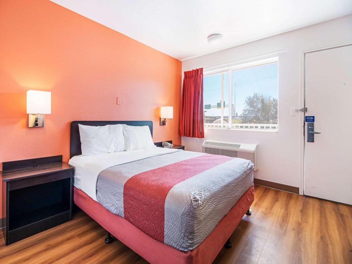 A bright room features an orange accent wall and natural light from a window. A spacious bed with crisp linens is positioned against the wall. A nightstand and air conditioning unit are visible, along with a wooden floor adding to the room's overall clean appearance.