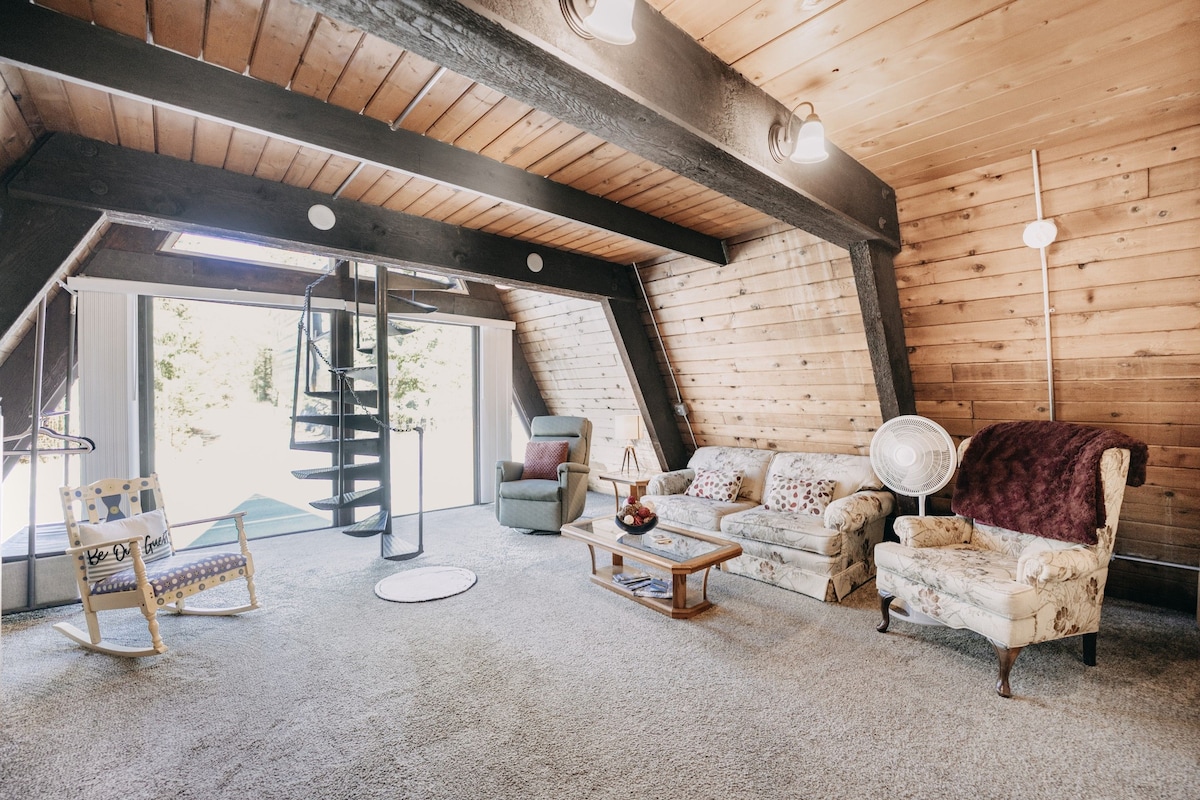 A spacious living area is highlighted by natural wood paneling and large windows providing ample natural light. Comfortable seating options, including a floral-patterned couch and a recliner, are accompanied by a coffee table. A spiral staircase leads to the loft bedroom, enhancing the open layout.