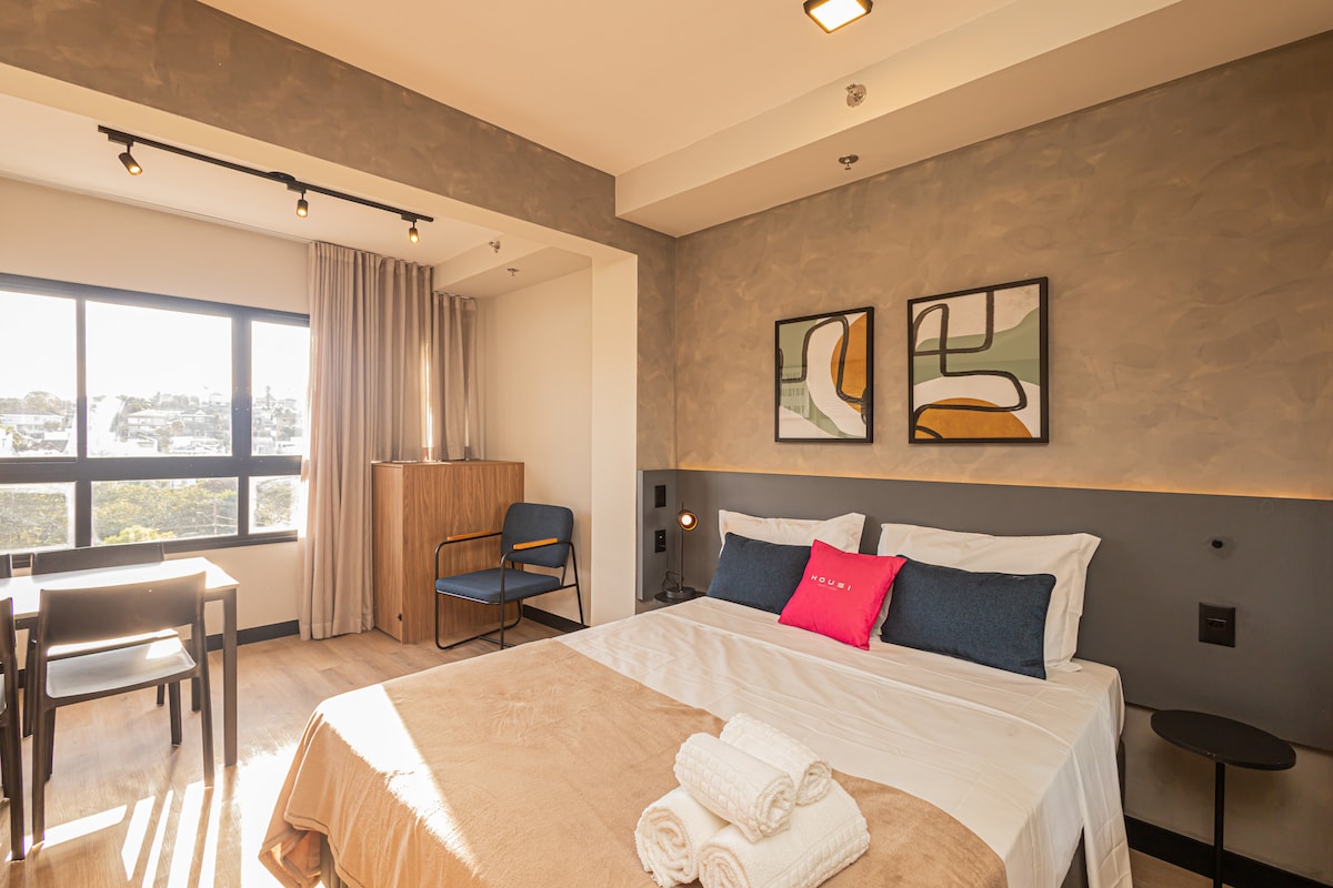 A bright room features a spacious bed adorned with plush pillows, complemented by a light blanket. Two framed artworks hang on the wall above the bed. A small dining table with two chairs is positioned near a window, which offers a view of the outdoors.