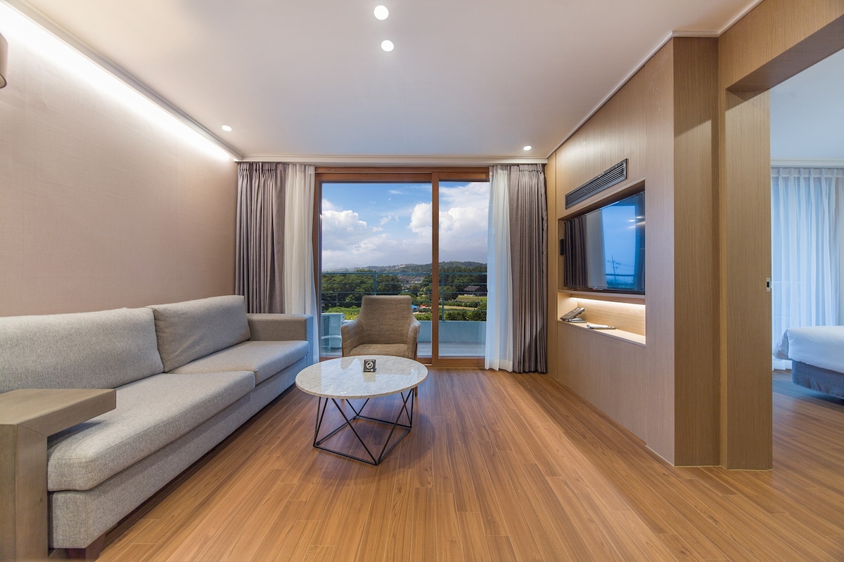 A modern living area features a light gray sofa positioned next to a geometric coffee table. Large windows offer scenic views of a lush landscape, allowing ample natural light to fill the space. A wall-mounted television is also visible, complementing the warm wood flooring.