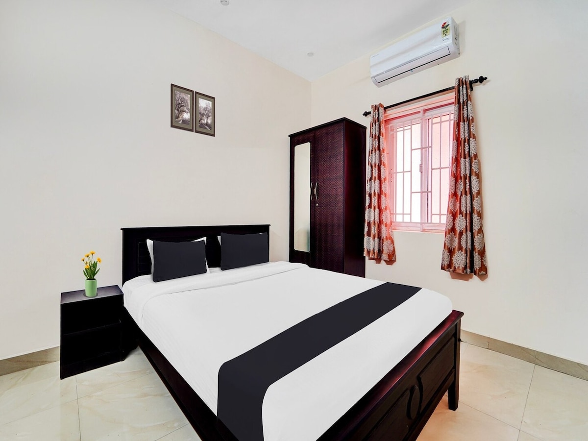 A well-lit bedroom features a spacious bed with dark bedding, complemented by matching pillows. A bedside table is positioned next to the bed, and a wardrobe with a mirrored door is visible. Natural light enters through a window framed by patterned curtains.