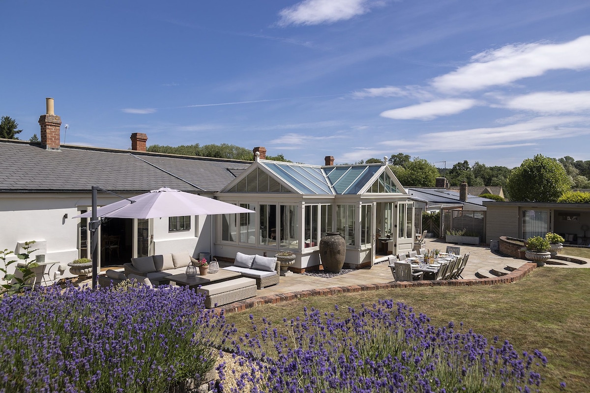 The exterior of the holiday cottage is displayed, showcasing a large conservatory with numerous windows, allowing ample light. The garden features a landscaped area with blooming lavender, multiple seating arrangements, and a shaded patio under a large umbrella.