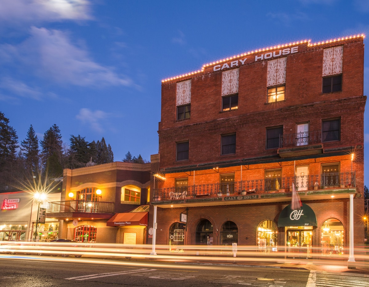 The historic Cary House Hotel is featured with its brick façade and illuminated signage. Warm lighting accentuates the building's architectural details, while nearby shops and streets are visible under a dusk sky. A sense of charm is evoked by the bustling atmosphere.