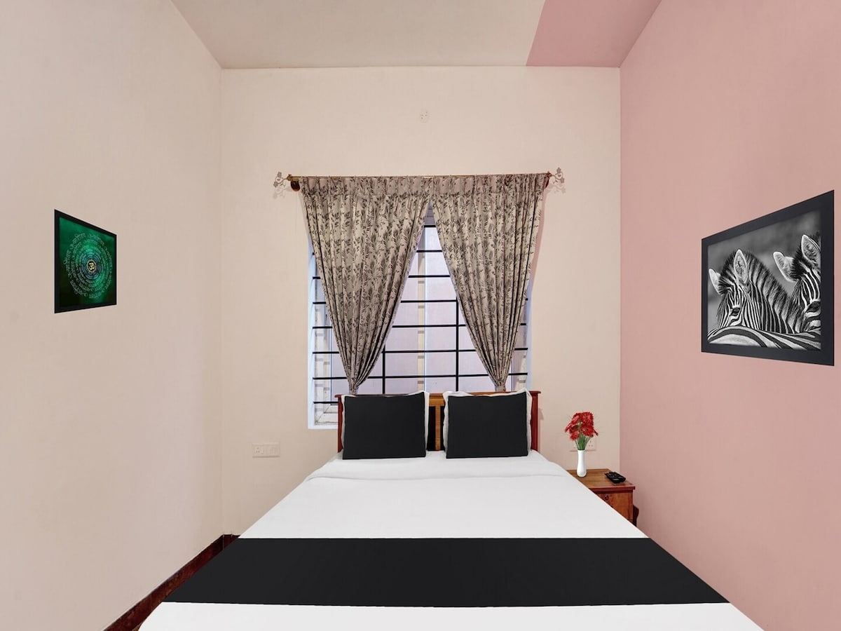 A well-organized bedroom features a spacious bed with a black and white striped cover. Two decorative paintings are mounted on the walls, complemented by light-filtering curtains at the window. A bedside table holds a small flower vase, adding a touch of color to the room.
