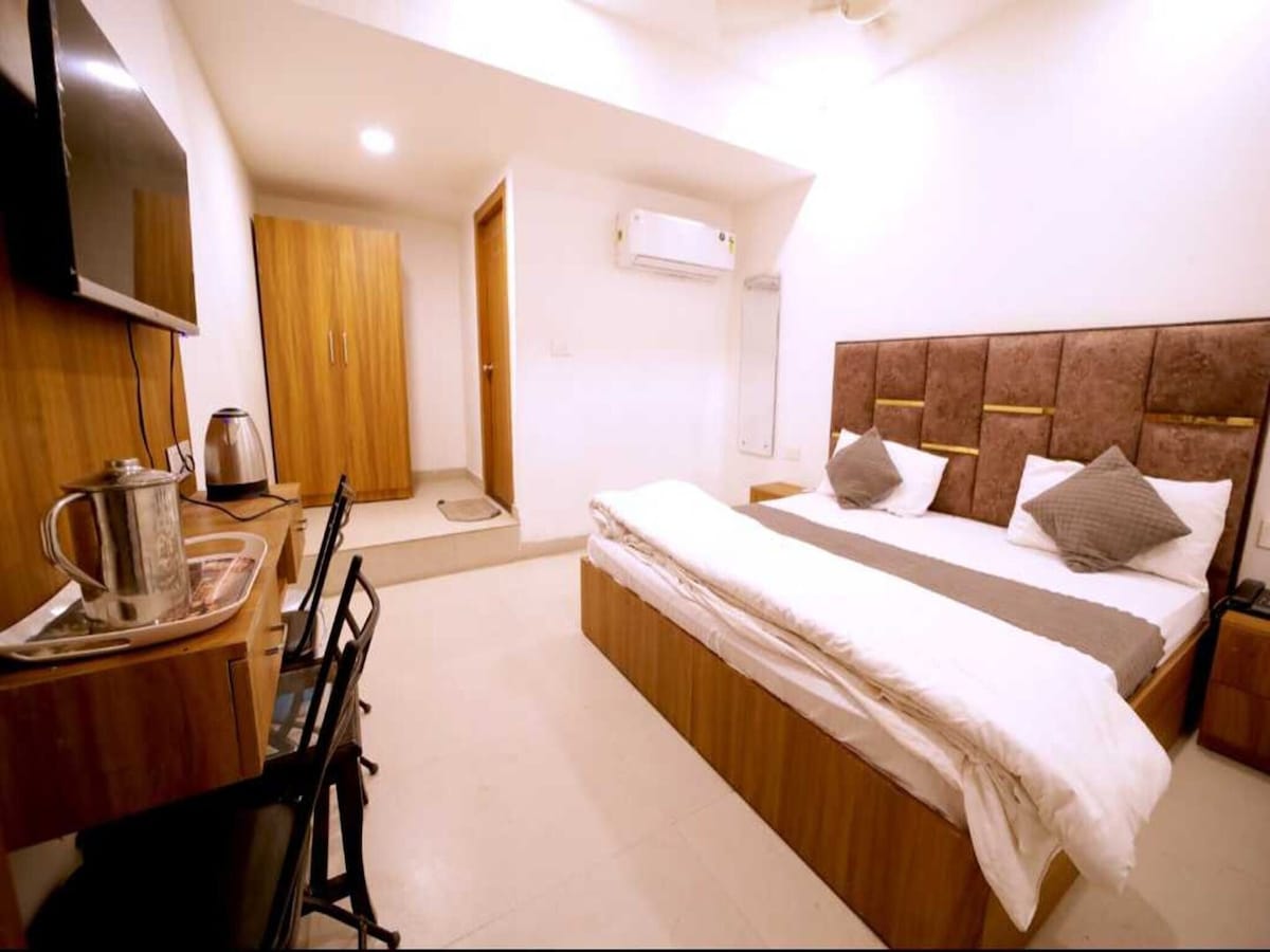 The guest room features a spacious bed with neutral bedding and decorative pillows, complemented by a wooden bedside table. A flat-screen TV is mounted on the wall opposite. A small seating area with a desk and chair is present, along with a wardrobe and a kettle on a tray.