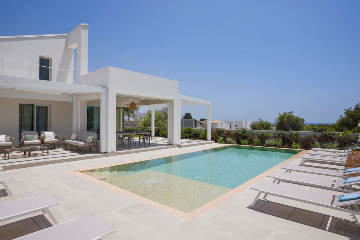 A modern villa exterior is presented, featuring a heated saltwater pool and a spacious sun terrace lined with loungers. Light-colored furniture is arranged around the pool, with a covered outdoor dining area visible, enhancing the connection between indoor and outdoor spaces.
