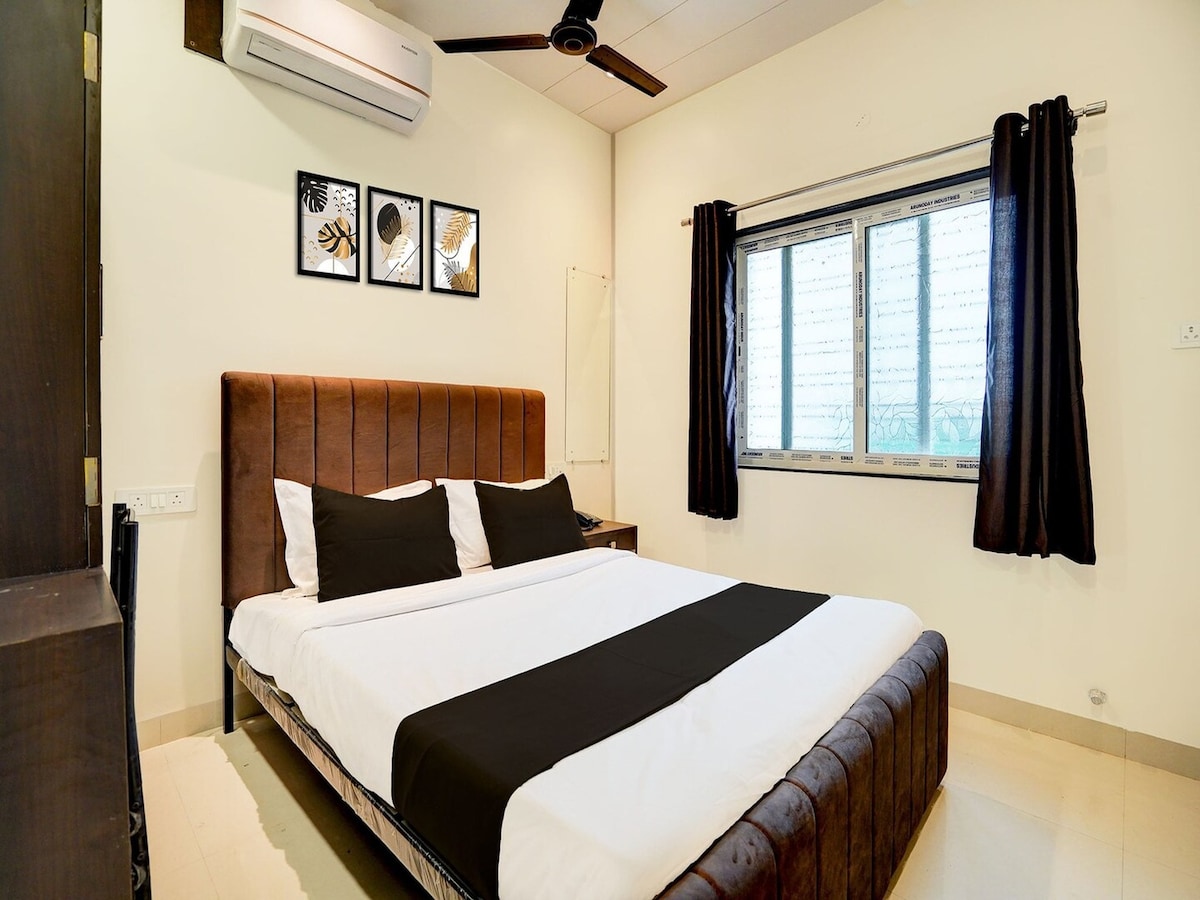 A spacious sleeping area features a large bed with dark linens, complemented by light-colored walls. Three framed artwork pieces hang above the headboard. A window covered with sheer curtains allows natural light, and a ceiling fan adds to the overall comfort.