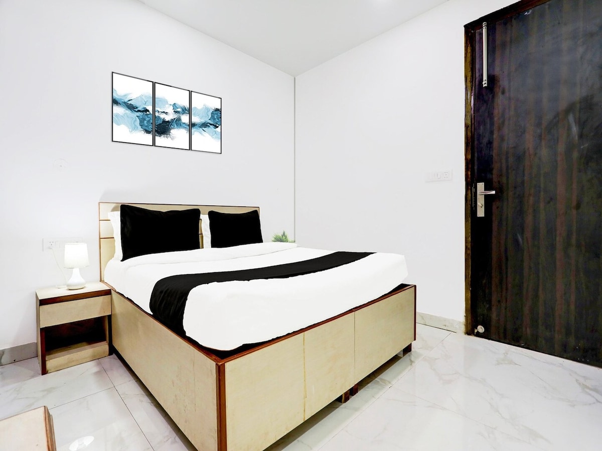 A well-appointed bedroom features a spacious bed covered with crisp white linens and a contrasting black runner. Minimalist artwork adorns the wall above the bed, while a bedside table holds a lamp. The room is complemented by white walls and a dark wooden door.