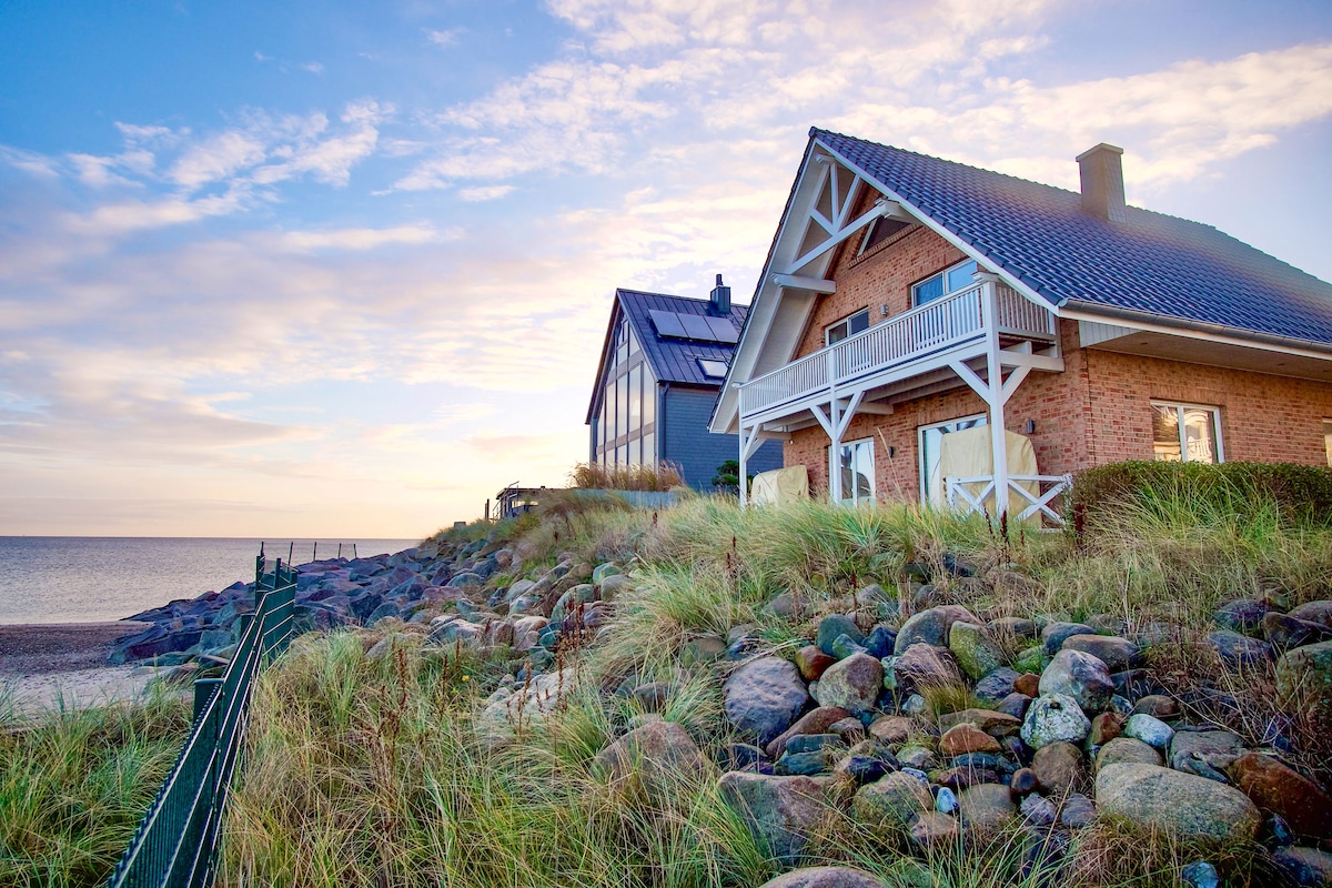 A beachfront holiday house is captured at sunset, showcasing its inviting architecture and the surrounding natural landscape. The blue sea is visible in the background, along with grassy dunes leading to the rocky shore, emphasizing the tranquility of the seaside setting.