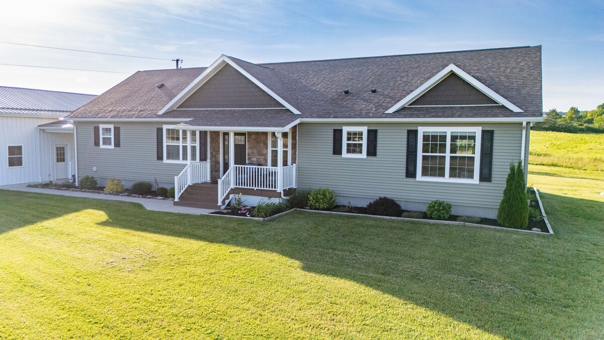 The exterior of the home features a well-maintained facade with a welcoming front porch and multiple windows maximizing natural light. A spacious lawn surrounds the house, complemented by neatly arranged shrubs and a panoramic view of the expansive landscape.