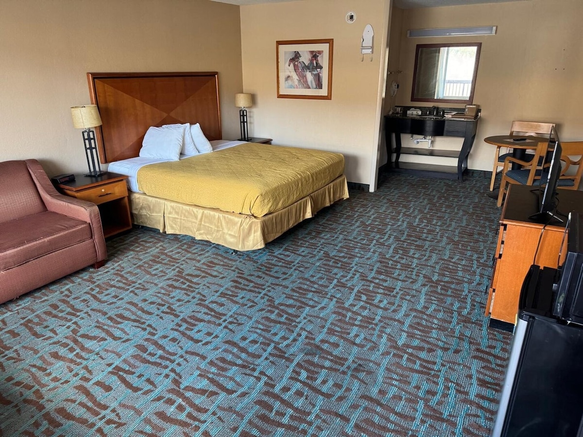 The room features a spacious king bed with a yellow bedspread and a wooden headboard. A red sofa sits adjacent to the bed, and a small dining table is set against the wall. A flat-screen TV and a desk with a chair are visible, along with a small fridge.
