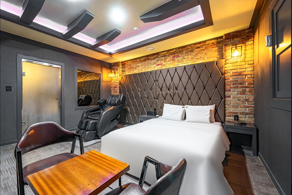 A modern room features a spacious bed with crisp white bedding, against a backdrop of textured brick and padded wall accents. A dark wood table and chairs provide a cozy seating area. Soft lighting and contemporary ceiling details enhance the room's overall ambiance.
