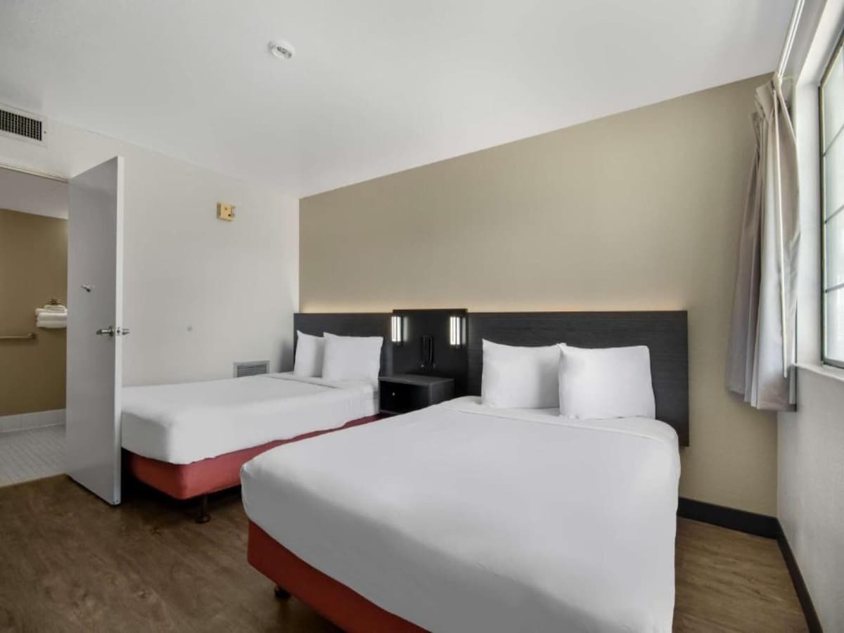 A guestroom features two spacious queen beds with light bedding, positioned against a wall with softly lit accents. A nightstand sits between the beds. Natural light enters through a nearby window with light curtains, illuminating the warm wood flooring.