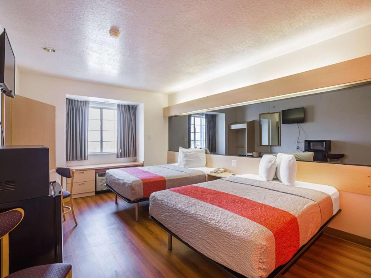 The guest room features two spacious beds with a simple grey and red bedspread. A large window allows natural light to enter, while an air conditioning unit is positioned beneath it. A flat-screen TV is mounted opposite the beds, and a desk area is visible in the room.